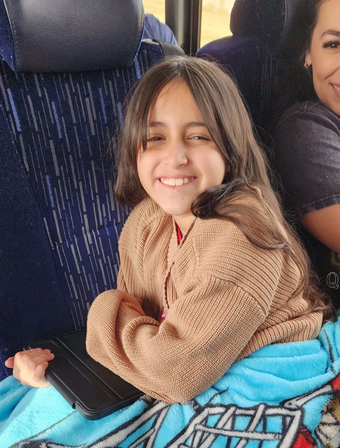 Two girls sitting on a bus with a blanket on their laps.